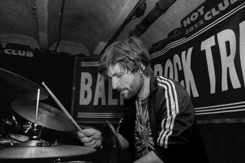 Black-and-white side-view of a drummter Todor Akimov in a striped jacket, head bowed in concentration as he grips a drumstick above his kit, with a “Rock Trio” banner and “Hot Jazz Club” sign visible behind him.
