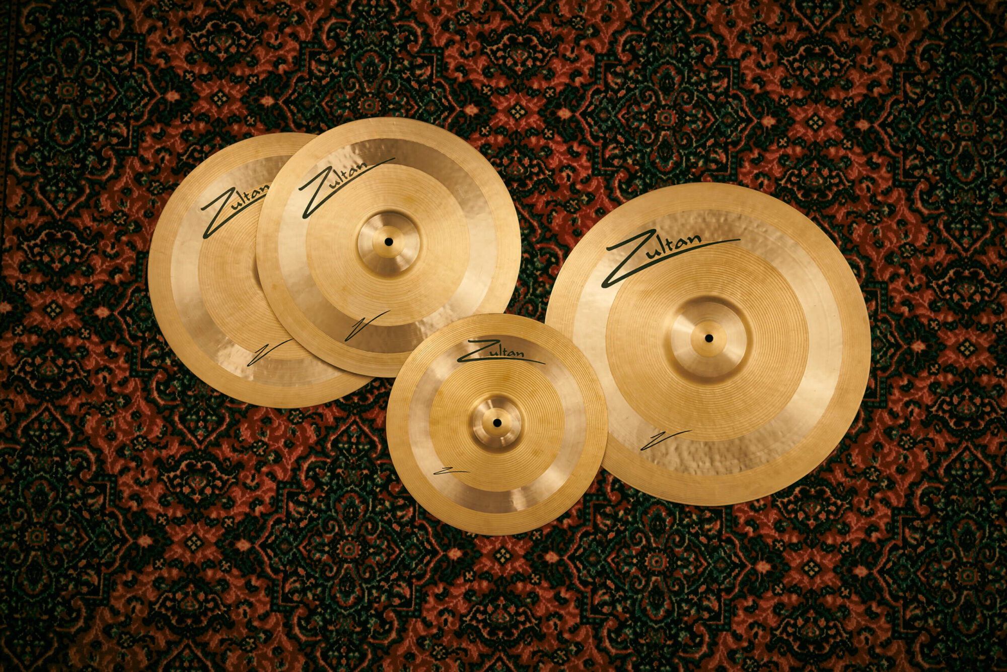 Overhead view of four polished bronze Zultan cymbals arranged on a red-and-green patterned rug: two mid-sized cymbals overlapping on the left, a large cymbal on the right, and a smaller splash cymbal at the bottom center, each bearing the stylized “Zultan” logo and signature mark.