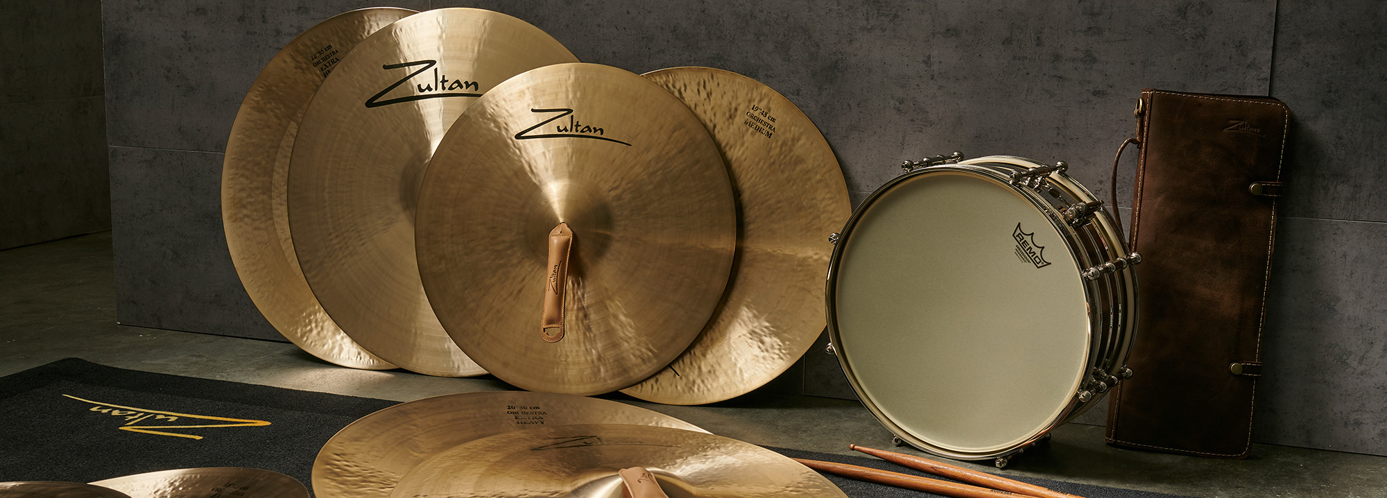 A collection of Zultan cymbals, a snare drum, a drumstick bag, and drumsticks arranged on a dark mat against a textured gray wall.
